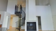 This two-storey space makes a strong architectural statement through its soaring double-height ceilings, which create an expansive, airy atmosphere in the main living area. The centerpiece of the space is a sleek, built-in fireplace framed by a clean white surround, adding both warmth and a refined focal point to the room. Positioned alongside it is a bold black metal spiral staircase, which serves as both a functional connector between the two floors and a striking design element in its own right. The staircase's curved form contrasts elegantly with the crisp, white walls that envelop the interior. Upper-level windows with louvred or slatted coverings allow natural light to filter into the space while maintaining privacy, illuminating the generous vertical volume. The second floor features a landing with matching black metal railings that echo the staircase's aesthetic, ensuring a cohesive design language throughout. The light hardwood flooring grounds the space with warmth, complementing the otherwise neutral, modern palette. Overall, the space delivers a sophisticated, loft-inspired feel with clean lines and thoughtfully integrated architectural details.
