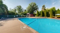 Located at 181 Nutmeg Ln in East Hartford, Connecticut, this outdoor recreational amenity area offers a well-appointed retreat for residents and guests. At the heart of the space is a generously sized freeform swimming pool with clear, bright blue water, featuring clearly marked depth indicators and designated no-diving zones for safety. The pool is surrounded by a broad, smooth concrete sundeck that provides ample room for relaxation and movement.

The deck is furnished with numerous lounge chairs and side tables, arranged to take full advantage of the open sky and natural sunlight. Several shade umbrellas in light blue are positioned throughout the area, offering comfortable spots to unwind out of the sun. A compact brick utility or pool house structure sits poolside, providing additional functionality to the space.

Adjacent to the pool area, a set of tennis courts enclosed by chain-link fencing offers additional recreational opportunities. The courts are accessible and well-maintained, with additional lounge seating positioned nearby for spectators. A green privacy-slated fence lines portions of the perimeter, enhancing seclusion and safety.

The entire area is framed by lush mature trees, including a row of tall evergreen arborvitae, creating a natural green backdrop that enhances the resort-like ambiance. The combination of aquatic and sport facilities, generous lounge space, and natural surroundings makes this a comprehensive outdoor amenity destination.