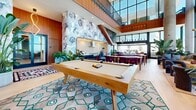 Located at 1401 Union St in San Diego's vibrant Bankers Hill neighborhood, this rooftop amenity level delivers an impressive blend of indoor sophistication and outdoor entertaining. Floor-to-ceiling glass walls flood the interior with natural light while framing sweeping panoramic views of the San Diego skyline and surrounding cityscape.

Inside, the lounge area is anchored by a curved, tufted caramel leather sectional sofa set against an expansive window wall, creating a dramatic focal point. Warm wood paneling lines the walls alongside a bold, large-scale floral wallcovering that adds an artistic flair. A large flat-screen display, shuffleboard table, and a full-sized pool table make this an ideal space for social gatherings and recreation. Intimate seating arrangements with leather lounge chairs and a marble coffee table round out the interior experience.

The outdoor terrace extends the living space significantly, featuring wicker lounge furniture, multiple seating vantage points, and a fully equipped outdoor kitchen with dual grilling stations and bar-height seating. Lush potted greenery and layered area rugs add warmth and texture to the tiled outdoor floor. Whether relaxing under open skies or entertaining guests, this elevated amenity space offers a seamless indoor-outdoor lifestyle with unobstructed views in every direction.