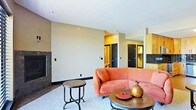 This single-floor space centers around a generously sized living area defined by its warm, neutral-toned walls and smooth white ceiling. The room's most striking feature is a curved terracotta sectional sofa, which anchors the space with a bold, retro-inspired silhouette. Paired with a round wrought-iron side table and a low circular coffee table adorned with decorative ceramic vessels, the seating arrangement creates an inviting and cohesive focal point.

A dark, marble-surround fireplace adds an elegant architectural element along one wall, contributing both visual depth and a sense of warmth to the room. The light-toned carpet softens the overall aesthetic and complements the earthy color palette throughout.

In the background, doorways lead to additional rooms, hinting at a well-connected interior layout. Wood-toned cabinetry is partially visible, suggesting built-in storage or a kitchen area nearby. Black-framed doors provide a subtle contrast to the warm wall tones, lending a contemporary edge to the overall design. The space balances comfort and style through its thoughtful material choices and cohesive warm color scheme.