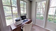 This single-floor space showcases a thoughtfully designed home office that blends classic architectural character with a warm, functional aesthetic. The office is defined by its striking herringbone-patterned brick flooring, which lends a timeless, traditional quality to the room. Large, multi-pane windows wrap around the space on two sides, flooding the interior with abundant natural light and framing picturesque views of mature trees and lush greenery beyond. The tall windows feature clean white millwork and paneled detailing that complement the room's refined styling. Subtle vertical wall paneling adds architectural depth and a polished finish to the interior. A rich walnut-toned wooden desk with brass hardware anchors the workspace, accompanied by a classic upholstered chair in neutral tones. The combination of natural materials, generous daylighting, and verdant outdoor views creates a serene and inspiring environment well-suited for focused work. The overall ambiance is one of quiet sophistication — grounded by natural textures, brightened by sunlight, and connected to the outdoors through its expansive window arrangement.
