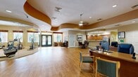 Located at 200 78th Ave N in Saint Petersburg, Florida, this community amenity center offers a thoughtfully designed single-floor layout encompassing a generous array of shared facilities. The welcoming reception and leasing office area greets visitors with warm hardwood-style flooring, a distinctive wave-pattern tray ceiling accented in rich golden-brown tones, recessed lighting, and dual workstations — creating a professional yet inviting atmosphere. Double glass entry doors allow natural light to flood the reception area, offering views of the lush Florida landscaping outside. The kitchen features rich wood cabinetry, granite countertops, stainless steel appliances including a full-size refrigerator, and tile flooring, complemented by pendant lighting and an open pass-through to a dining area with a glass-top table. The expansive exercise room is a standout feature, equipped with treadmills, ellipticals, stationary bikes, and strength-training machines, all set on plush carpet with floor-to-ceiling glass walls and mirrored surfaces that enhance the sense of space. A dedicated computer station area provides individual wooden workstations for residents. Connecting hallways with hardwood-style floors and neutral walls tie all areas together seamlessly, creating a cohesive and functional community hub.