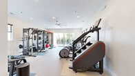 Located at 12027 Saint Vrain Rd in Longmont, Colorado, this fitness center is a comprehensively outfitted single-room facility designed to support a full range of workout routines. The space is bright and clean, featuring white walls, recessed lighting, and ceiling fans that maintain a comfortable exercise environment. Large sliding glass doors and multiple windows along the perimeter flood the room with natural light while offering views of the surrounding greenery, and a connected outdoor deck provides additional ventilation and openness. The flooring combines carpet and rubberized mat areas, delineating zones for different types of exercise. Cardio equipment includes treadmills, stair climbers, elliptical trainers, and stationary bikes, all positioned to take advantage of the natural light and outdoor views. A dedicated strength training zone features a multi-function cable and pulley machine, a full rack of dumbbells with an adjustable bench, and a wall-mounted functional training rig stocked with resistance bands, medicine balls, and stability balls. The layout is thoughtfully organized to maximize usable floor space while keeping traffic flow clear between stations, making this facility well-suited for diverse fitness needs.