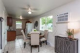 This single-floor home offers a thoughtfully organized layout with a variety of functional spaces connected by light tile flooring throughout the main areas. The kitchen is designed for efficiency, featuring rich dark-countered cabinetry lining both walls, a window overlooking the backyard, and ample storage. It opens to a casual dining area furnished with a round wooden table and tufted upholstered chairs, brightened by large windows that bring in generous natural light and views of the surrounding greenery. A ceiling fan and wall-mounted air conditioning unit provide year-round comfort in this space. The living room is generously sized, centered around a large sectional sofa with tile flooring, a ceiling fan, and a window framing neighborhood views. A carpeted bedroom offers a peaceful retreat with a ceiling fan, window with blinds, and baseboard heating. A separate carpeted room serves as a functional home office, complete with a ceiling fan, window, and baseboard heating. The two bathrooms are finished with light tile flooring, dark-countered wood vanities, and well-lit mirrors — one featuring a tub-shower combo and the other a standalone soaking tub. Clean white walls and consistent finishes lend a cohesive, move-in-ready feel throughout the home.