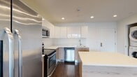 Located at 10 DeKalb Ave in White Plains, NY, this modern one-bedroom, one-bathroom residence offers a thoughtfully designed open-concept layout with clean lines and contemporary finishes throughout. Rich dark hardwood flooring flows seamlessly across the living, dining, and bedroom areas, providing a warm contrast to the bright white walls and recessed lighting overhead.

The kitchen is a standout feature, equipped with high-gloss white upper cabinetry, natural wood-toned lower cabinets, and light quartz countertops. A central island with a matching countertop provides additional prep and dining space. Stainless steel appliances — including a range, microwave, dishwasher, and refrigerator — complete the well-appointed culinary space.

The open living area is bright and airy, anchored by floor-to-ceiling windows that invite abundant natural light and offer views of the surrounding White Plains cityscape. The bedroom continues the dark hardwood flooring and features a dedicated closet for ample storage.

The bathroom is finished with large-format floor tiles, a wall-mounted vanity, frameless mirror, and a full tub and shower with decorative tile accents. A stacked in-unit washer and dryer add everyday convenience to this well-designed residence.