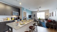 This thoughtfully designed single-floor space showcases a seamless open-concept layout that connects the kitchen, dining, and living areas into one cohesive and functional environment. The kitchen is outfitted with stainless steel appliances, including a range, microwave, and ample cabinetry in a rich dark finish. A granite-topped breakfast bar provides casual seating for multiple guests and serves as a natural divider between the kitchen and living spaces. Track lighting overhead ensures the kitchen is well-lit and practical. A teal accent wall adds a bold pop of color, anchoring the living area where a flat-screen TV, open shelving, and comfortable seating create an inviting atmosphere. A ceiling fan enhances comfort in the living space, which benefits from a large window that floods the room with natural light. The light-toned flooring flows consistently throughout, contributing to a clean, airy feel. A glimpse into an adjacent room suggests additional living or sleeping quarters, further extending the functionality of the space. The overall ambiance is modern and welcoming, blending neutral tones with intentional color accents for a balanced, contemporary aesthetic.