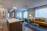 This single-floor residence showcases a contemporary design aesthetic defined by clean lines, warm wood tones, and a carefully curated mix of light and dark finishes throughout. The open-concept kitchen, dining, and living area forms the social heart of the home. The kitchen is outfitted with two-tone cabinetry combining white upper cabinets and warm walnut-toned lower cabinets, stainless steel appliances, a white quartz countertop island with bar seating, subway tile backsplash, a wine cooler, and pendant lighting overhead. Light hardwood flooring extends through the main living area, which is bathed in natural light through large windows dressed with patterned curtains and white wood blinds. The living room features a bold yellow sofa, a wall-mounted TV against a deep slate-blue accent wall, and a ceiling fan. The adjacent dining area offers additional seating and flows seamlessly into the kitchen. The bedrooms continue the sophisticated palette, with dark charcoal accent walls, ceiling fans, plush carpeting, and large windows allowing ample natural light. The bathroom presents a walnut vanity with white quartz countertops, an illuminated mirror, and a tiled shower enclosure, completing the residence's polished, modern character.