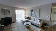 This single-floor space offers a clean, contemporary aesthetic highlighted by a well-appointed living area. The living room serves as the focal point, bathed in natural light streaming through expansive sliding glass doors dressed with vertical blinds that offer both privacy and brightness control. The room features a plush, light gray sectional sofa accented with decorative pillows, positioned over a geometric patterned area rug that adds texture and visual interest to the space. A round, metallic coffee table anchors the seating arrangement, complementing the modern sensibility of the room. A large abstract canvas artwork in muted tones of gray, white, and gold adorns the main wall, adding a sophisticated artistic touch. The neutral gray walls and textured ceiling create a calm, inviting ambiance throughout. Warm accent lighting from a floor lamp enhances the cozy atmosphere, especially during evening hours. The sliding glass doors provide a seamless connection to the outdoors, allowing ample natural light to fill the space. The overall design embraces a cohesive neutral palette with carefully chosen furnishings that balance comfort and style, making this a welcoming and versatile living environment.
