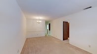 This single-floor space in Summit presents a straightforward and functional layout with a warm, neutral aesthetic throughout. The main living area is finished with wall-to-wall beige carpeting that extends seamlessly into adjoining spaces, creating a cohesive and comfortable feel underfoot. Walls are painted in a soft, off-white tone, lending the interior a clean and airy atmosphere that serves as a versatile backdrop for a variety of furnishings and décor styles. A traditional chandelier with candelabra-style arms serves as a subtle focal point overhead, adding a touch of classic charm to the space. A decorative chair rail runs along the wall, providing a modest architectural detail that adds visual interest. Doorways connect to additional rooms, suggesting a practical and well-organized floor plan. Natural light filters in from an adjoining room visible in the background, contributing to the overall brightness of the interior. The space presents a blank canvas, well-suited for those looking to move in and make it their own.