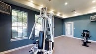 This single-level fitness room offers a thoughtfully designed workout space finished with a distinctive slate-blue color palette across its walls, complemented by crisp white baseboards and trim. The ceiling features a recessed tray design with abundant recessed lighting, ensuring the space is well-lit throughout. A ceiling fan provides additional air circulation during workouts.

The room is carpeted throughout, providing a comfortable surface underfoot. It is equipped with a multi-station cable weight machine for comprehensive strength training, along with a treadmill, elliptical trainer, recumbent bike, and an upright stationary bike for varied cardiovascular exercise options. A wall-mounted television is positioned for viewing during workouts.

Multiple windows along the walls allow natural light to filter in, offering views of lush greenery outside and creating an open, airy atmosphere. A wall-mounted mirror near the strength equipment provides visual feedback during exercise. A glass-paneled entry door adds a bright, welcoming touch to the space. Overall, the room strikes a balance between functional fitness amenities and an inviting, clean aesthetic.