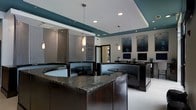 This single-floor space presents a contemporary and polished interior designed to make a strong visual impression. The focal point of the room is a substantial granite-topped counter with rich, dark wood cabinetry beneath, providing both a functional workspace and an elegant design element. A coffered ceiling with teal-painted recessed sections adds depth and color contrast against the white upper portions, while a series of pendant lights hang at varying heights, contributing to the sophisticated ambiance.

The space features curved booth-style seating with dark upholstery, arranged to create distinct zones within the open layout. A decorative chainmail-style partition screen adds a unique textural element, serving as both a visual divider and an artistic feature. Recessed lighting supplements the pendant fixtures, ensuring the space is well-illuminated throughout.

Along the back wall, wall-mounted screens and framed artwork provide visual interest, while the curved reception-style counter creates a natural flow through the room. The neutral carpeted flooring complements the darker furnishings, and a glass door at the rear hints at additional spaces beyond. Overall, the design balances function with contemporary style.