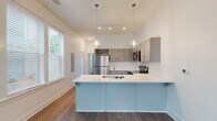 This contemporary two-story home in Warner Robins, Georgia offers a well-designed layout with modern finishes throughout. The main level centers around an open-concept living and kitchen area, where durable wood-look flooring, crisp white walls, and an abundance of natural light from large windows create a bright and welcoming atmosphere. The kitchen is a standout feature, showcasing a distinctive light-blue peninsula with a white quartz countertop and an integrated sink, complemented by gray shaker-style cabinetry, white tile backsplash, and a full suite of stainless steel appliances including a refrigerator, range, microwave, and dishwasher. Pendant lighting and a track light fixture add a stylish touch overhead. A staircase with dark wood treads and a warm wood handrail leads to the upper level, where the bedrooms are located. Each bedroom offers wood-look flooring, ample closet space, and access to natural light. The bathrooms are clean and functional, each featuring a tub-shower combination, white vanity with quartz countertop, gray cabinetry, and wood-look flooring. A dedicated laundry area with a full-size washer and dryer adds everyday convenience. The home's cohesive design and quality finishes make it a comfortable and practical residence.