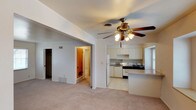 This single-floor residence offers a practical and functional open-concept layout that seamlessly connects the main living area to the kitchen and beyond. The living room is finished with wall-to-wall carpeting in a neutral beige tone, creating a warm and comfortable foundation. A ceiling fan with an integrated light fixture is centrally mounted, providing both air circulation and ambient lighting to the space.

The kitchen is positioned adjacent to the living area and is accessible via an open pass-through design, featuring white cabinetry that extends to the upper walls, a refrigerator, a range with hood, and a raised countertop that serves as a natural dividing bar between the two spaces. The kitchen floor transitions to a light-toned tile, distinguishing the functional cooking zone from the carpeted living area.

A wide arched entryway leads from the living room into a hallway, offering access to additional rooms including what appears to be a closet and a bathroom with a warm wood-toned door. The walls throughout are painted in soft, neutral shades of white and light gray, contributing to a clean and airy ambiance. Recessed ventilation fixtures are present throughout the ceiling, reflecting a thoughtfully designed interior layout suited for comfortable everyday living.