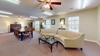 Located at 391 Old Griffin Rd in McDonough, Georgia, this single-floor community space offers a warm and functional layout designed for gathering and socializing. The main open area seamlessly combines a lounge and dining zone, carpeted throughout and bathed in natural light from multiple large windows fitted with blinds. The lounge is anchored by a curved sectional sofa, a glass-topped coffee table, and a ceiling fan with integrated lighting, creating a comfortable and inviting atmosphere. Framed artwork and table lamps add a refined touch to the warm, neutral-toned walls.

The dining section accommodates a long table with ample seating, making it well-suited for group meals or activities. A dedicated piano nook features an upright piano, a wooden bookcase, and additional seating, offering a quiet corner for leisure. A large mirror and black accent chairs line one wall near the entryway, providing a welcoming transition into the main space. Tall indoor plants are placed throughout, adding a natural, lively element to the interior.

The kitchen is a practical galley-style space with honey-toned wood cabinetry, laminate countertops, and a striking black-and-white checkered tile floor. It is equipped with a full-size refrigerator, electric range, microwave, and sink, supporting full catering or event needs. A utility corridor connects the kitchen to the main gathering area efficiently.