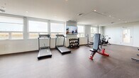 Located at 2453 Renfrew St in Vancouver, BC, this dedicated fitness facility offers a thoughtfully designed exercise space across a single, open floor. The room is finished with a durable rubber flooring surface and clean white walls, creating a bright and functional environment conducive to focused workouts.

The space is well-lit by a combination of recessed ceiling lights and generous natural light streaming through large windows along one wall, which also frame pleasant outdoor views. A full-length wall mirror spans one end of the room, visually expanding the space and allowing users to monitor their form during exercise.

The facility is equipped with a diverse range of fitness equipment to support both cardio and strength training. Cardio options include multiple treadmills and a rowing machine, while strength training is supported by a power rack loaded with weight plates, a adjustable bench, a dumbbell rack with a full set of free weights, and stationary air bikes. A mounted flat-screen TV provides entertainment or guided workout content during sessions. A built-in drinking fountain on one wall adds a practical convenience for users.

The layout is open and well-organized, allowing for comfortable movement between equipment zones. With its modern finishes, abundant natural light, and comprehensive equipment selection, this exercise room delivers a complete and inviting fitness experience.
