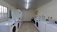 This shared laundry facility is a practical, single-floor space designed to accommodate high-volume laundry needs. The room is outfitted with multiple front-loading washing machines and stacked dryer units arranged along both sides, maximizing the available floor space and allowing several users to run laundry simultaneously. The units are arranged in an organized, efficient layout that promotes easy access and smooth circulation throughout the room. A utility sink is positioned along one wall, providing a convenient station for hand-washing or pre-treating garments. The flooring consists of durable stone-effect tiles that are well-suited for a high-traffic utility environment. Overhead fluorescent lighting ensures the room is brightly and evenly illuminated. The walls are finished in a clean, neutral tone, contributing to a tidy and functional atmosphere. A corkboard is mounted on one wall, serving as a notice board for posted information or community announcements. The overall design of the space prioritizes practicality and efficiency, providing a clean and accessible laundry environment suited for shared residential or commercial use.