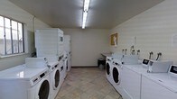 This shared laundry facility is a practical, single-floor space designed to accommodate high-volume laundry needs. The room is outfitted with multiple front-loading washing machines and stacked dryer units arranged along both sides, maximizing the available floor space and allowing several users to run laundry simultaneously. The units are arranged in an organized, efficient layout that promotes easy access and smooth circulation throughout the room. A utility sink is positioned along one wall, providing a convenient station for hand-washing or pre-treating garments. The flooring consists of durable stone-effect tiles that are well-suited for a high-traffic utility environment. Overhead fluorescent lighting ensures the room is brightly and evenly illuminated. The walls are finished in a clean, neutral tone, contributing to a tidy and functional atmosphere. A corkboard is mounted on one wall, serving as a notice board for posted information or community announcements. The overall design of the space prioritizes practicality and efficiency, providing a clean and accessible laundry environment suited for shared residential or commercial use.