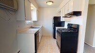 This updated one-bedroom, one-bathroom Canterbury unit is thoughtfully laid out across a single floor, offering a functional and comfortable living environment. The galley-style kitchen is efficiently designed with white cabinetry lining both walls, light countertops, a stainless double-basin sink, and black appliances including a gas range, refrigerator, and dishwasher. Light tile flooring and a ceiling fixture keep the space bright and easy to maintain. The open living area is generously proportioned, featuring plush carpeting, warm neutral-toned walls, and multiple windows that invite abundant natural light and outdoor views of the surrounding brick building exterior. A ceiling fan with light fixture, wall-mounted air conditioning unit, and baseboard heating ensure year-round comfort. The bedroom continues the carpeted flooring and neutral palette, with a large window framing outdoor views and ample natural light, accompanied by closet access and baseboard heating. The bathroom is well-appointed with wood-look flooring, a white vanity with a large mirror, a tub and shower combination, a window for natural ventilation, and classic tile wainscoting. The overall layout, as reflected in the dollhouse view, is compact yet efficient, making excellent use of the available space.