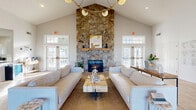 Located at 6601 Oak Hill Circle in Rensselaer, NY, this single-floor community clubhouse offers a thoughtfully designed gathering space that blends rustic charm with modern comfort. The centerpiece of the space is a grand lounge area anchored by a soaring vaulted ceiling and a dramatic floor-to-ceiling stone fireplace with a reclaimed wood mantel. A contemporary multi-globe chandelier hangs above, casting warm light over two plush neutral sofas flanking a live-edge coffee table atop a woven area rug. Light wood-tone flooring and an abundance of natural light from multiple French doors and transom windows create an airy, inviting atmosphere throughout. Adjacent to the lounge, a dedicated leasing office provides a clean, professional setting with a large white desk and guest seating beneath high ceilings. The fitness center is well-appointed with cardio equipment including treadmills and a stationary bike, as well as a multi-functional strength training station, free weights, and exercise accessories, all set on durable rubber flooring. A bright, functional kitchen rounds out the space, featuring white shaker cabinetry, gray quartz countertops, stainless steel appliances, and recessed lighting — ideal for community events and gatherings.
