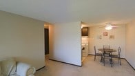 This single-floor residence features an open-concept layout that seamlessly connects the living, dining, and kitchen areas into one cohesive space. The living area is finished with wall-to-wall carpeting in a neutral beige tone, contributing to a warm and understated aesthetic. A textured popcorn ceiling runs throughout, and the walls are painted in a soft, neutral palette that keeps the space feeling bright and consistent.

A partial dividing wall separates the living area from the kitchen zone, providing a subtle sense of definition without fully closing off the space. The kitchen is equipped with white appliances, including a freestanding electric range, and is complemented by dark cabinetry above, offering a mild contrast to the lighter surroundings. The kitchen benefits from its own overhead lighting.

Adjacent to the kitchen, a modest dining area accommodates a small table and chairs, making it a functional spot for everyday meals. A hallway visible from the main living space leads toward additional rooms, suggesting a practical and straightforward floor plan. Overall, the space presents a clean, functional layout suited for comfortable everyday living.
