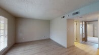 This single-story unit offers a straightforward and functional layout across several distinct areas. The living room is a generously proportioned space finished with light hardwood-style flooring and neutral-toned walls, complemented by a textured popcorn ceiling. A large window allows natural light to fill the room, while a front entry door provides direct access to the space. HVAC vents are visibly integrated into the ceiling, ensuring climate control throughout.

Adjacent to the living area, a secondary room features the same light hardwood-style flooring and connects to a hallway corridor leading further into the unit. The kitchen is equipped with dark espresso-toned cabinetry, a double stainless steel sink, and granite-patterned countertops, providing ample workspace and storage. Light hardwood-style flooring continues in this area.

The bedroom is carpeted and benefits from a window with a blind, admitting natural light, along with a ceiling-mounted light fixture. It connects directly to the bathroom, which features a dark wood vanity with a granite-topped countertop, a framed mirror, a tub-shower combination, and tile surround. Clean, neutral finishes are consistent throughout, giving the unit a cohesive and understated aesthetic.