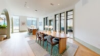 Located at 4101 Raleigh St in Charlotte, NC, this single-floor space offers a striking blend of architectural character and functional design. The layout is anchored by a series of elegant arched openings that frame intimate built-in booth seating, each alcove finished with vertical wood slat paneling, curved LED accent lighting, and lush greenery — creating cozy, semi-private nooks within the larger open plan. The main area features wide-plank light oak hardwood floors that run throughout, complementing the soft sage green accents found on walls, door frames, and architectural arches. Two generously sized communal tables — one with a distinctive scalloped edge — occupy the central gathering zone, adorned with globe table lamps and surrounded by a mix of upholstered and wooden chairs. A patterned green-and-white checkered area rug grounds the seating clusters and adds a curated, design-forward touch. Floor-to-ceiling windows on one end flood the space with natural light while offering views of the surrounding streetscape. Artful wall hangings and a carefully selected furniture palette give the space a warm, editorial quality. Recessed ceiling lighting supplements the abundant natural light, ensuring the space remains bright and welcoming throughout the day.