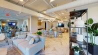 Located at 1150 E Street in downtown San Diego, this single-floor amenity space combines modern industrial design with refined, contemporary furnishings across several distinct zones. Exposed ductwork and track lighting run along the ceilings throughout, lending an open, loft-inspired aesthetic to the entire floor.

The main lounge area features stunning marble-patterned flooring, an elegant chandelier, and a large flat-screen TV, surrounded by plush sofas and stylish seating arrangements. Decorative shelving, lush indoor plants, and curated art pieces — including striking woven wall hangings — enhance the space's warm, welcoming ambiance.

A café-style dining area with round tables and contemporary chairs opens up to floor-to-ceiling windows, offering abundant natural light and views of surrounding palm trees and the urban San Diego skyline. A secondary lounge area is finished with neutral carpet, modular seating, circular accent mirrors, and intimate bistro tables, ideal for casual gatherings or focused work.

A dedicated entertainment lounge includes a large mounted television and comfortable sectional seating, perfect for gaming or media viewing. A glass-walled conference room provides a sophisticated setting for meetings, complete with an elegant chandelier, a long wood table, and upholstered office chairs, all visible through sliding glass doors.