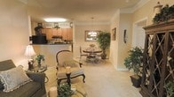 This single-floor residence exudes a warm, traditional elegance throughout its open and thoughtfully arranged layout. The living area anchors the space with plush carpeting, classic upholstered seating, and tasteful decorative touches including lush greenery and candle accents that contribute to a refined, inviting ambiance. Crown molding and a chair rail lend architectural character to the walls, reinforcing the timeless design sensibility found throughout.

Flowing seamlessly from the living area, the dining space features a wrought-iron and glass dining table set beneath a decorative chandelier, adding a touch of sophistication. A large framed mirror on the wall enhances the sense of depth and brightness in the dining area, while an elegant indoor tree adds a natural, organic element to the room.

The kitchen is positioned just beyond, accessible via a raised breakfast bar that offers a convenient transition between cooking and entertaining spaces. Warm honey-toned cabinetry extends to the ceiling, maximizing storage, while black appliances provide a subtle contrast. The overall color palette throughout the space is soft and neutral, with cream and beige tones creating a cohesive, calming atmosphere. This residence balances comfort with classic style in a well-connected, single-level floor plan.