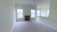 This single-floor residential space offers a clean, well-lit interior designed for comfortable everyday living. The main living area is a standout feature, characterized by plush wall-to-wall carpeting and a generously proportioned layout that provides ample room for furnishing and movement. A tile-surround fireplace serves as the central focal point of the room, flanked by multiple windows that invite an abundance of natural light throughout the day. Crown molding along the ceiling adds a refined architectural detail, lending a polished feel to the overall aesthetic. The neutral, light-toned walls complement the soft carpeting and create a warm, welcoming ambiance throughout the space. Track lighting is installed near the ceiling, providing flexible, directed illumination to supplement the natural light. A partial view of an adjacent area — likely a kitchen or dining space — is visible, hinted at by a dark countertop edge, suggesting a practical and connected layout. The windows, positioned on multiple walls, offer views of the surrounding exterior environment and further enhance the sense of openness. Overall, the space presents a tidy, move-in ready interior with classic residential finishes suitable for a variety of living arrangements.