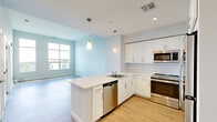 Located at 7 Crown Dr in Quincy, MA, this thoughtfully designed one-bedroom, one-bathroom residence offers a clean, modern aesthetic across a single floor. The open-concept kitchen and living area serves as the heart of the home, featuring light hardwood-style flooring, soft gray walls, and abundant natural light streaming through large windows. The kitchen is well-appointed with white flat-front cabinetry, light stone countertops, stainless steel appliances including a refrigerator, range, microwave, and dishwasher, and a pendant light accent — creating a bright and functional cooking space. The living area flows seamlessly from the kitchen, providing a versatile layout with access to the bedroom and bathroom through crisp white panel doors. The bedroom is a comfortable retreat finished with plush carpeting, a large window overlooking the surrounding neighborhood, and soft neutral walls. The bathroom features wood-look tile flooring in warm tones, offering a clean and practical space. Throughout the residence, consistent light gray walls, white trim, and coordinated flooring choices create a cohesive, contemporary atmosphere that is both inviting and easy to personalize.