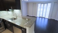 This single-floor space showcases a thoughtfully designed open-concept layout that seamlessly connects the kitchen and living area. The kitchen is a standout feature, equipped with dark espresso cabinetry that contrasts sharply against a light granite countertop with rich veining. A peninsula with a built-in sink and stainless steel fixtures extends into the space, offering additional prep area and a natural boundary between the kitchen and living zone. A decorative mosaic tile backsplash in neutral tones adds texture and visual interest to the kitchen walls. Stainless steel appliances, including a dishwasher, are integrated beneath the counter for a streamlined look. The flooring throughout is rich, dark hardwood, lending warmth and continuity across both the kitchen and living areas. The living space is generously sized and benefits from an abundance of natural light streaming through floor-to-ceiling windows dressed with horizontal blinds. A subtle column detail and clean crown molding add architectural refinement. The overall ambiance is modern and polished, with a neutral color palette that creates a bright, welcoming environment.