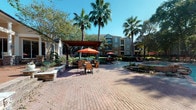 This outdoor amenity space serves as a central gathering hub within a well-maintained residential community. The layout is anchored by a generously sized swimming pool with clearly marked depth zones and a smooth paver pool deck lined with blue lounge chairs, providing ample space for sunbathing and relaxation. Adjacent to the pool, a charming pergola structure offers a shaded retreat, complemented by wicker dining tables and chairs topped with vibrant orange umbrellas — ideal for casual outdoor dining. Additional seating areas with umbrella-covered tables are thoughtfully distributed across the expansive brick-paved courtyard, creating comfortable spots for socializing throughout the day. Lush, manicured landscaping including tall palms, ornamental trees, trimmed hedgerows, and seasonal plantings adds greenery and visual interest throughout the space. The surrounding multi-story buildings feature classic brick facades with bay windows, balconies, and pitched rooflines that lend a traditional architectural character to the community. Mature trees provide natural shade across walkways and seating nodes, enhancing comfort in the outdoor environment. The overall ambiance is inviting and resort-like, combining practical amenities with attractive landscaping to create a vibrant outdoor living experience.