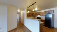 This single-floor space showcases a well-designed open layout that seamlessly connects the kitchen to the adjoining living areas. The kitchen is a central highlight, featuring warm honey-toned wood cabinetry that extends to the upper walls, providing generous storage. A peninsula-style breakfast bar with a granite-look countertop serves as both a workspace and a casual dining surface, creating a natural divide between the kitchen and the rest of the space.

Overhead, a black track lighting rail paired with pendant lights in warm tones ensures ample task lighting throughout the kitchen, while flush-mount ceiling fixtures illuminate the hallway and adjacent areas. Black appliances, including a range, add a sleek contrast to the lighter cabinetry and countertops.

The flooring throughout is a warm, medium-toned hardwood-style material that adds continuity and cohesion across the open floor plan. A hallway visible beyond the kitchen leads to additional rooms, suggesting a practical and well-organized layout. The neutral wall tones and warm lighting contribute to an inviting, comfortable ambiance throughout the space.