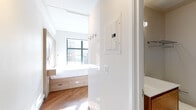 This thoughtfully designed urban studio showcases a clean, modern aesthetic throughout its single-floor layout. The sleeping area is anchored by a platform bed with built-in drawer storage and a warm natural wood headboard wall panel, complemented by a mounted reading lamp and rich hardwood flooring that runs throughout the space. Large floor-to-ceiling windows flood the room with abundant natural light while framing expansive city skyline views, creating a striking urban backdrop. A built-in window bench with understated storage adds both function and charm to the living area, which also features a compact dining table and wall-mounted television. The kitchenette is efficiently laid out, featuring light wood cabinetry, white upper cabinets, a vibrant turquoise glass backsplash, an induction cooktop, stainless steel microwave, and a full-size refrigerator — all within a compact but well-appointed footprint. The bathroom is finished with large-format tile flooring, a floating wood-toned vanity, and a frameless glass walk-in shower, offering a refined and spa-like feel. Track lighting and pendant fixtures contribute to the space's modern, urban character throughout.