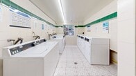 Located at 79 Jameson Avenue in Toronto's Parkdale neighbourhood, this shared laundry facility is a well-organized, functional space designed to serve building residents efficiently. The room is laid out with rows of top-loading and front-loading washing machines along both walls, complemented by a lineup of dryers, providing ample capacity for multiple users. A utility sink with exposed faucet fixtures is available for hand-washing or pre-treatment needs. The interior features white ceramic wall tiles accented by a distinctive green tile border running along the upper portion of the walls, lending a clean and classic institutional aesthetic. Underfoot, patterned vinyl floor tiles in a neutral geometric design add a subtle visual element while remaining practical and easy to maintain. Overhead, linear fluorescent lighting ensures the space is brightly and evenly illuminated. A window at the far end of the room allows for natural light and a street-level view of the surrounding neighbourhood. A built-in countertop provides a convenient folding and sorting surface, and a cork-board tenant notification board is mounted on the wall, keeping residents informed of building updates and laundry instructions. The space is tidy, well-maintained, and clearly organized for ease of use.