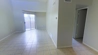 This single-floor space offers a clean, functional layout designed to maximize openness and comfort. The main living area is finished with light-toned ceramic tile flooring that extends throughout, creating a cohesive and easy-to-maintain surface underfoot. Notably high, vaulted ceilings elevate the sense of space, allowing the interior to feel expansive and breathable.

Natural light flows generously into the living area through a full-width sliding glass door fitted with vertical blinds, which opens directly onto a balcony. This outdoor access provides views of the surrounding environment and adds an inviting indoor-outdoor connection to the space.

The walls are finished in a neutral, light gray tone that complements the pale tile flooring, resulting in a bright and understated palette throughout. HVAC vents are positioned both in the ceiling and along the lower walls, ensuring consistent climate control across the space. A partial wall divides the living area, providing subtle architectural definition without compromising the open feel of the floor plan.

Overall, the space presents a straightforward, well-lit interior that balances practicality with an airy, comfortable ambiance, well-suited for a variety of uses and easily adaptable to different furnishing styles.
