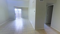 This single-floor space offers a clean, functional layout designed to maximize openness and comfort. The main living area is finished with light-toned ceramic tile flooring that extends throughout, creating a cohesive and easy-to-maintain surface underfoot. Notably high, vaulted ceilings elevate the sense of space, allowing the interior to feel expansive and breathable.

Natural light flows generously into the living area through a full-width sliding glass door fitted with vertical blinds, which opens directly onto a balcony. This outdoor access provides views of the surrounding environment and adds an inviting indoor-outdoor connection to the space.

The walls are finished in a neutral, light gray tone that complements the pale tile flooring, resulting in a bright and understated palette throughout. HVAC vents are positioned both in the ceiling and along the lower walls, ensuring consistent climate control across the space. A partial wall divides the living area, providing subtle architectural definition without compromising the open feel of the floor plan.

Overall, the space presents a straightforward, well-lit interior that balances practicality with an airy, comfortable ambiance, well-suited for a variety of uses and easily adaptable to different furnishing styles.