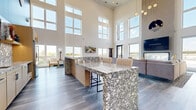 Located at 710 Vikings Parkway in Eagan, Minnesota, this impressive single-floor amenity lounge is designed to deliver a premium communal experience. The space is immediately defined by its dramatic double-height ceilings and an extensive array of floor-to-ceiling windows that flood the interior with natural light and offer sweeping outdoor views of the surrounding landscape.

The open-plan layout flows seamlessly from one functional zone to the next, all unified by stylish wide-plank hardwood-style flooring. A standout kitchen and bar area anchors one end of the space, featuring a striking granite-patterned stone countertop island with modern bar seating, light wood cabinetry, open display shelving, and a stainless steel French door refrigerator — ideal for gatherings and events.

The main lounge area is generously appointed with a billiards table, comfortable seating arrangements, a fireplace, and multiple wall-mounted televisions, creating an inviting atmosphere for socializing or relaxing. A dedicated games area includes a regulation shuffleboard table, set alongside sleek metal railings and modern wall sconces that add a polished, contemporary aesthetic. Pendant lighting and carefully chosen décor elements complete the sophisticated, modern design throughout this versatile communal space.