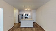 This single-floor space showcases a thoughtfully designed open-concept layout, with the kitchen serving as a central highlight. The kitchen is outfitted with stainless steel appliances, including a refrigerator, microwave, and range, all set against crisp white cabinetry and a classic subway tile backsplash. A substantial island with granite countertops provides additional workspace and serves as a natural gathering point, illuminated by elegant glass pendant lights suspended overhead. A track lighting fixture adds supplemental task lighting to the cooking area. The rich, dark hardwood-style flooring runs throughout, creating a warm contrast against the light neutral walls and white cabinetry. The open layout allows the kitchen to flow seamlessly into the surrounding living areas, contributing to an airy and spacious feel. Clean white baseboards and paneled doors add a polished, finished look to the interior. The overall design aesthetic is contemporary and clean, combining functional kitchen elements with modern finishes to create a cohesive and inviting living environment.
