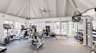 This fitness center is set within a single-story building distinguished by its striking vaulted, cathedral-style ceiling with white-painted exposed beams and shiplap detailing. A ceiling fan is centrally mounted at the peak, providing air circulation throughout the space. The bright, airy atmosphere is enhanced by an abundance of large, multi-pane windows and French doors lining the walls, offering views of the surrounding exterior and flooding the interior with natural light.

The flooring throughout is durable rubber matting, well-suited for a fitness environment. The space is comprehensively equipped for both cardiovascular and strength training. Cardio offerings include treadmills, an elliptical, and a stationary cycle. Strength training is supported by a multi-station cable machine, a full rack of dumbbells, adjustable benches, and various selectorized weight machines. Stability balls and a wall-mounted flat-screen television round out the amenities.

Full-length mirrors are positioned along one wall, visually expanding the space while serving a practical function for exercise form. The overall design aesthetic is clean and bright, with white walls and trim creating a cohesive, polished look throughout.
