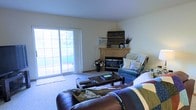 This single-floor space centers around a well-appointed living room designed for comfort and functionality. The room features a corner fireplace with a light wood mantel, serving as a natural focal point and decorative anchor. Above the mantel, rustic wooden décor adds a warm, homey character to the space. The living area is carpeted and generously sized, accommodating a substantial leather seating arrangement alongside a wooden coffee table, creating a welcoming gathering space. A large sliding glass door along one wall floods the room with natural light and provides views of an outdoor area, enhancing the sense of openness. The neutral-toned walls complement the varied textures found throughout the room, from the plush carpet underfoot to the mix of upholstered and leather furniture. A teal armchair positioned near the fireplace adds a pop of color, while decorative floral accents and throw pillows contribute to the overall warmth of the interior. The layout is practical and open, making efficient use of the available space while maintaining a comfortable, residential atmosphere throughout.