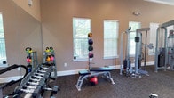 This fitness center offers a practical and well-organized workout environment designed to support a variety of exercise routines. The space is finished with warm beige walls and durable carpet flooring, creating a comfortable atmosphere for training. Natural light filters in through multiple windows fitted with horizontal blinds, keeping the space bright and inviting. A large mirror along one wall enhances the sense of openness and allows users to monitor their form during workouts. The facility is stocked with an impressive range of equipment, including a full rack of dumbbells in varying weights, a set of colorful kettlebells on a dedicated stand, and a medicine ball tower holding multiple weighted balls. A versatile adjustable workout bench sits at the center of the space, ideal for a wide range of strength exercises. A multi-function cable resistance machine rounds out the equipment selection, providing options for upper and lower body training. The thoughtful layout ensures ample room to move freely between stations, making the space functional for both individual and simultaneous use. Overall, this fitness center delivers a clean, well-lit, and comprehensively equipped environment suited for strength training and functional fitness.