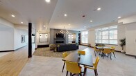This contemporary community lounge space is thoughtfully designed to offer a dynamic blend of comfort, style, and functionality across a single open floor. The layout is anchored by a striking floor-to-ceiling shiplap fireplace wall, complete with a mounted flat-screen television, creating a warm and inviting focal point in the main lounge area. Plush gray sofas and bold patterned accent chairs in yellow and black are arranged around a central coffee table atop a textured area rug, evoking a relaxed yet sophisticated atmosphere. An eye-catching geometric chandelier with exposed Edison-style bulbs hangs overhead, adding an artistic touch to the space. Adjacent to the lounge, multiple dining tables with vibrant yellow chairs offer flexible seating arrangements for gatherings. Large windows along one wall allow generous natural light to fill the space, enhancing its open and airy feel. The kitchenette and bar area is a standout feature, showcasing a dramatic dark marble-topped island with a decorative patterned base, sleek black pendant lighting, a geometric tile backsplash, stainless steel appliances, and open display shelving. Light-toned tile flooring flows throughout, tying together the space's modern, upscale aesthetic.
