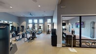 This modern fitness center is thoughtfully designed to support a full range of workout routines across multiple distinct zones. The main exercise area is expansive and airy, featuring soaring ceilings and a generous row of large windows that invite abundant natural light and views of the surrounding outdoor scenery. The space is finished with durable speckled flooring and painted in a contemporary cool gray palette throughout.

The cardio zone is well-stocked with multiple Matrix-brand treadmills, elliptical trainers, and other cardiovascular machines arranged along the windowed walls. A large wall-mounted television and floor-to-ceiling mirrors enhance the workout experience. The strength training section provides a selection of cable and resistance machines, complemented by ceiling fans that promote airflow and comfort.

A glass-walled enclosure separates a dedicated multi-purpose studio room, visible from the main floor, which houses additional specialty equipment including stability balls and resistance tools. Adjacent to this is a separate yoga and stretching studio with warm hardwood flooring, mirrored walls, a yoga mat, and direct access to the outdoors—creating a calm, focused environment for low-impact training.

Recessed lighting, hand sanitizer stations, and a ceiling fan complete the thoughtful amenities throughout this comprehensive fitness facility.