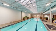 Located at 420 East Ohio Street in Chicago's Streeterville neighborhood, this expansive amenity space offers a comprehensive suite of recreational and leisure facilities across a single floor spanning a generously sized footprint. At its heart is a large indoor lap pool bathed in natural light from an overhead skylight ceiling, flanked by wood-plank pool decking and furnished with wicker lounge chairs. The pool area connects seamlessly to an outdoor terrace via large folding glass doors, where a barrel sauna and additional lounge seating provide a resort-like retreat. The outdoor spaces are thoughtfully divided into distinct zones: a composite wood deck lined with dark patio dining sets accommodates group gatherings, while lush artificial turf areas host recreational amenities including a ping-pong table and cornhole boards. String lighting overhead and vibrant botanical murals add character to the terrace. An outdoor kitchen station features stainless steel grills, a granite countertop bar, and barstool seating, ideal for al fresco entertaining. A separate courtyard area offers large communal dining tables set against the building's curved limestone façade. Surrounded by the Chicago skyline, this amenity space delivers a dynamic urban outdoor experience.