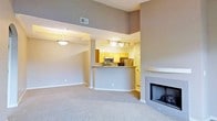 This single-floor space features an open-concept layout that seamlessly connects the living area and kitchen, creating a functional and inviting environment. The living room is carpeted in a neutral tone and anchored by a sleek fireplace with a clean, minimalist surround — an ideal focal point for the room. High ceilings with elevated upper walls enhance the sense of spaciousness, while a stylish pendant light fixture adds warmth to the dining or living area.

The kitchen is partially open to the main living space, separated by a breakfast bar that doubles as a casual dining or prep surface. Warm honey-toned cabinetry provides ample storage, complemented by stainless steel appliances including a microwave and refrigerator. Track lighting above the kitchen ensures the workspace is well-lit. A columned divider subtly defines the transition between the kitchen and living areas while maintaining the open feel.

Throughout the space, a consistent warm gray color palette on the walls ties each area together cohesively. The combination of soft carpet, warm cabinetry, and modern fixtures gives the space a comfortable yet polished ambiance suitable for everyday living.