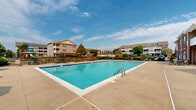 Located at 400 Haslet St in Dover, Delaware, this outdoor amenity area centers around a generously sized rectangular swimming pool with crystal-clear blue water, accessible via stainless steel entry rails. The pool is surrounded by a wide, open concrete sundeck that provides ample space for lounging, with multiple chaise lounge chairs and patio umbrellas positioned throughout for sun and shade options. Scattered seating arrangements invite relaxation and social gatherings in the open air.

The complex features a mix of architectural styles, with a classic red brick multi-story building accented by blue shutters flanking one side of the pool, and a neutral-toned, gabled clubhouse-style structure with white-trimmed windows and glass-panel doors situated directly adjacent to the pool deck. The clubhouse provides convenient access to interior facilities, and an HVAC unit is visible along the exterior wall.

Mature trees and well-maintained landscaping frame the perimeter of the community, offering a pleasant, park-like backdrop. A colorful playground with a slide is visible in the background, adding a family-friendly dimension to the outdoor space. The open sky and surrounding residential buildings contribute to a community-oriented atmosphere, making this outdoor area a central hub for recreation and leisure.