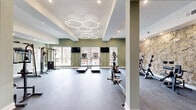 Located at 1818 N 53rd St in Philadelphia, PA, this fitness center offers a spacious, single-floor exercise room thoughtfully designed to support a wide range of workout routines. The space immediately impresses with its eye-catching honeycomb-patterned LED ceiling fixtures, which cast a bright, energizing glow throughout the room. Sage green walls provide a calm, motivating atmosphere, while a striking textured stone accent wall adds a bold architectural focal point and visual depth to the interior.

The exercise area is outfitted with a comprehensive selection of equipment, including a cable resistance machine, a power rack with barbells and weight plates, a dumbbell rack, a rowing machine, an elliptical trainer, treadmills, and a dedicated rack stocked with kettlebells and medicine balls. Rubber and carpet flooring throughout provide safety and comfort underfoot. Large windows along one wall flood the room with natural light and offer views of the surrounding neighborhood.

A wall-mounted flat-screen television is positioned for easy viewing during cardio sessions. Full-length mirrors on one wall visually expand the space and assist with proper form. Recessed lighting supplements the statement ceiling fixtures to ensure the room remains well-lit at all hours, making this a fully functional and attractively designed fitness facility.