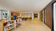 Located at 501 Broadway in Santa Monica, California, this thoughtfully designed lobby and communal space offers a sophisticated blend of functionality and contemporary style across a single floor. Large floor-to-ceiling windows along the exterior walls flood the interior with natural light and offer views of the surrounding streetscape, while sheer white curtains provide a soft, airy ambiance throughout.

The open-plan layout is divided into distinct functional zones. A generously sized lounge area anchors the space, furnished with curated mid-century modern seating, including accent chairs, a statement marble coffee table, and bold patterned area rugs that add visual warmth to the light tile flooring. A dedicated dining area features a solid wood table surrounded by classic cane-back chairs, set against a warm, illuminated wall display showcasing framed artwork.

Lush indoor plants are strategically placed throughout, enhancing the welcoming atmosphere with natural texture and greenery. Open wooden shelving units serve as stylish room dividers, adding depth and organization to the space.

A separate hallway area reveals a row of private meeting pods framed by dark metal and warm wood paneling, set against a deep navy accent wall — ideal for focused work or confidential conversations. Recessed ceiling lighting throughout ensures consistent, comfortable illumination across all zones.