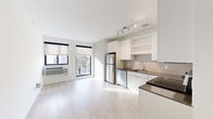 This single-floor unit in Minneapolis's 55406 zip code showcases a clean, modern design with an open-concept layout connecting the living area and kitchen. Light hardwood-style flooring runs throughout, lending a bright, cohesive feel to the space.

The kitchen is well-appointed with white flat-front cabinetry, open shelving, dark countertops, and a neutral tile backsplash. Stainless steel appliances include a refrigerator, electric range, microwave, and dishwasher, making the space both functional and contemporary.

The living area is filled with natural light through large, street-facing windows fitted with roller shades, offering views of the surrounding Minneapolis neighborhood. A wall-mounted HVAC unit provides climate control.

A hallway leads to the remaining areas of the unit, with built-in cabinetry providing generous storage along the corridor. A stacked in-unit washer and dryer is conveniently tucked within a dedicated closet along this hall.

The bathroom features light wood-style flooring consistent with the rest of the unit, a floating vanity with a white countertop and integrated sink, a large mirror, and a combined tub and shower. The overall palette of whites and neutrals creates a calm, minimalist ambiance throughout.